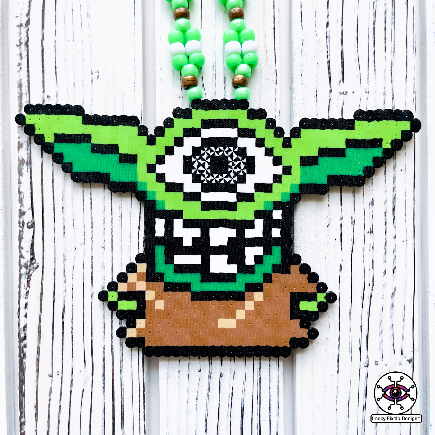 Baby grogu subtronics perler necklace. green yoda with cyclops eye and chaotic teeth. necklace is made with brown, white and green perler beads. He is wearing a brown outfit.