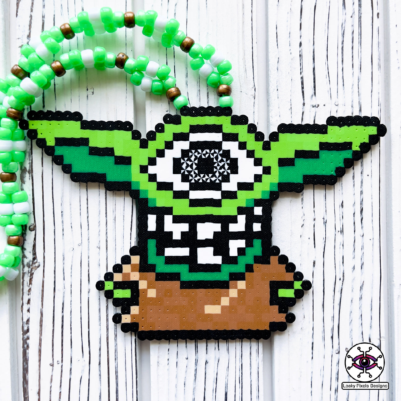 Baby grogu subtronics perler necklace. green yoda with cyclops eye and chaotic teeth. necklace is made with brown, white and green perler beads. He is wearing a brown outfit.