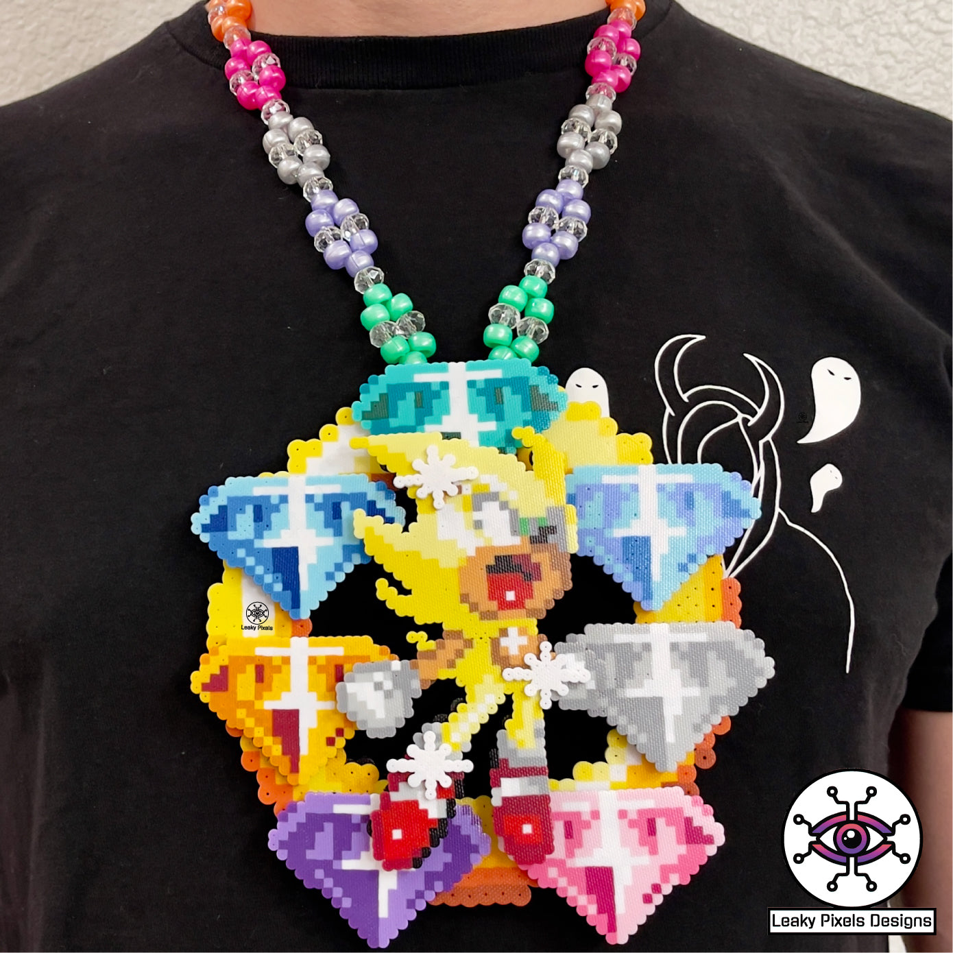 hyper sonic perler necklace by leaky pixels. yellow hyper sonic with stars around him, placed over 7 super emeralds green, blue, light blue, white, pink, orange and purple, on top of a gold ring. necklace is made of same colors of gems.