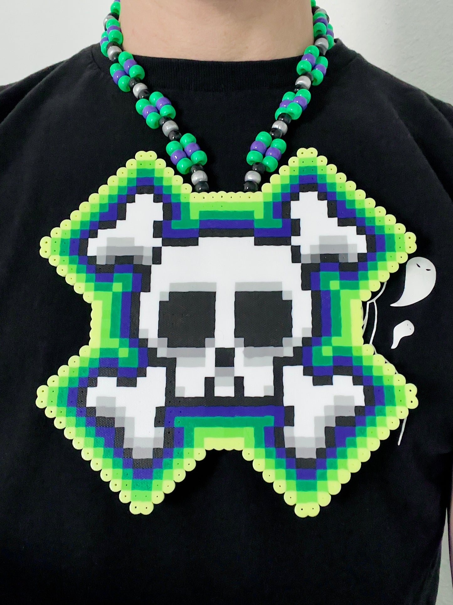 Skull and crossbones perler necklace by leaky pixels. skull and crossbones perler with green and purple colors radiating out of it. pony beads of green, purple, black and pearl silver make the necklace.