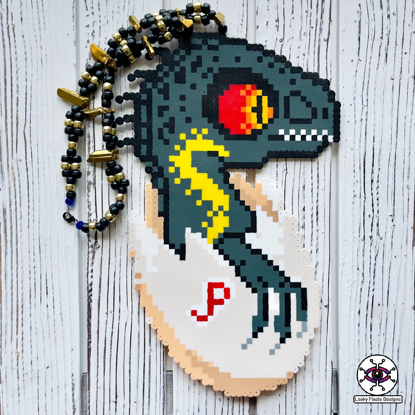 Indoraptor Baby in a half cracked egg shell with the letters JP inscribed onto the shell by leaky pixels. it is a perler necklace.