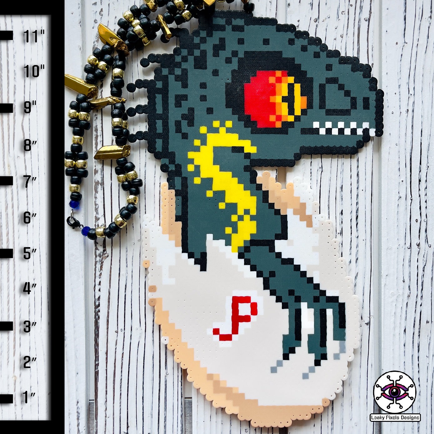 Indoraptor Baby in a half cracked egg shell with the letters JP inscribed onto the shell by leaky pixels. it is a perler necklace.