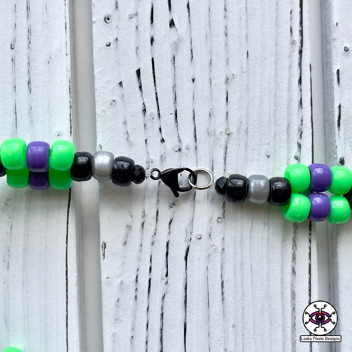 Skull and crossbones perler necklace by leaky pixels. skull and crossbones perler with green and purple colors radiating out of it. pony beads of green, purple, black and pearl silver make the necklace.