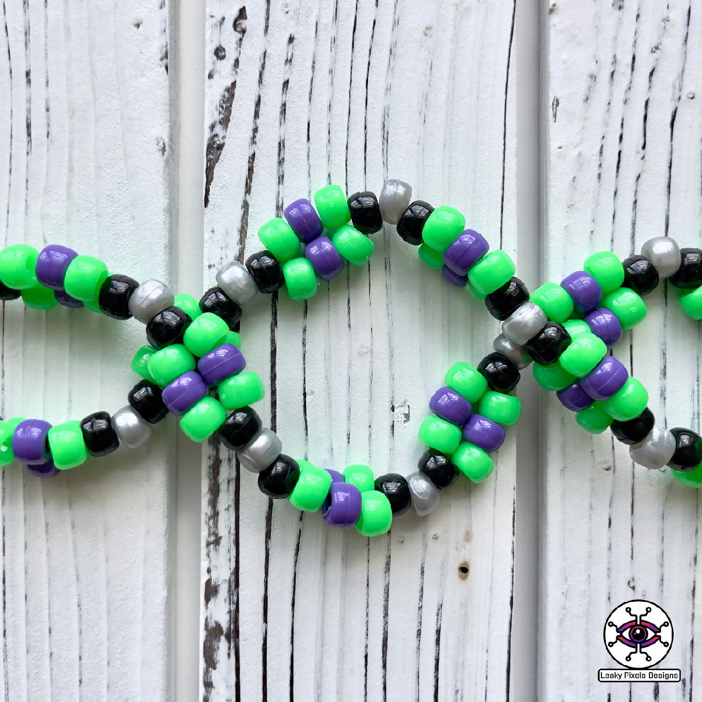 Skull and crossbones perler necklace by leaky pixels. skull and crossbones perler with green and purple colors radiating out of it. pony beads of green, purple, black and pearl silver make the necklace.