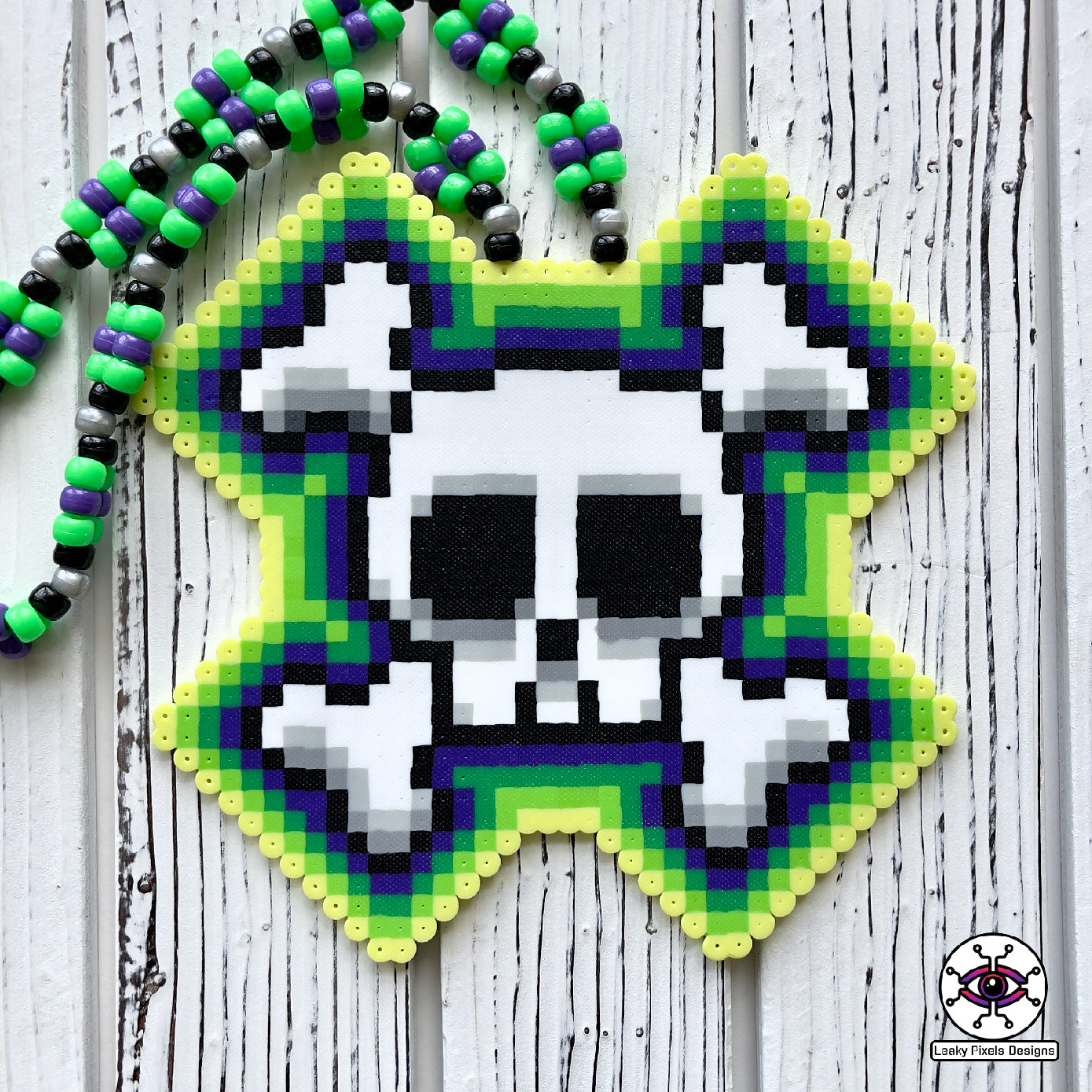 Skull and crossbones perler necklace by leaky pixels. skull and crossbones perler with green and purple colors radiating out of it. pony beads of green, purple, black and pearl silver make the necklace.