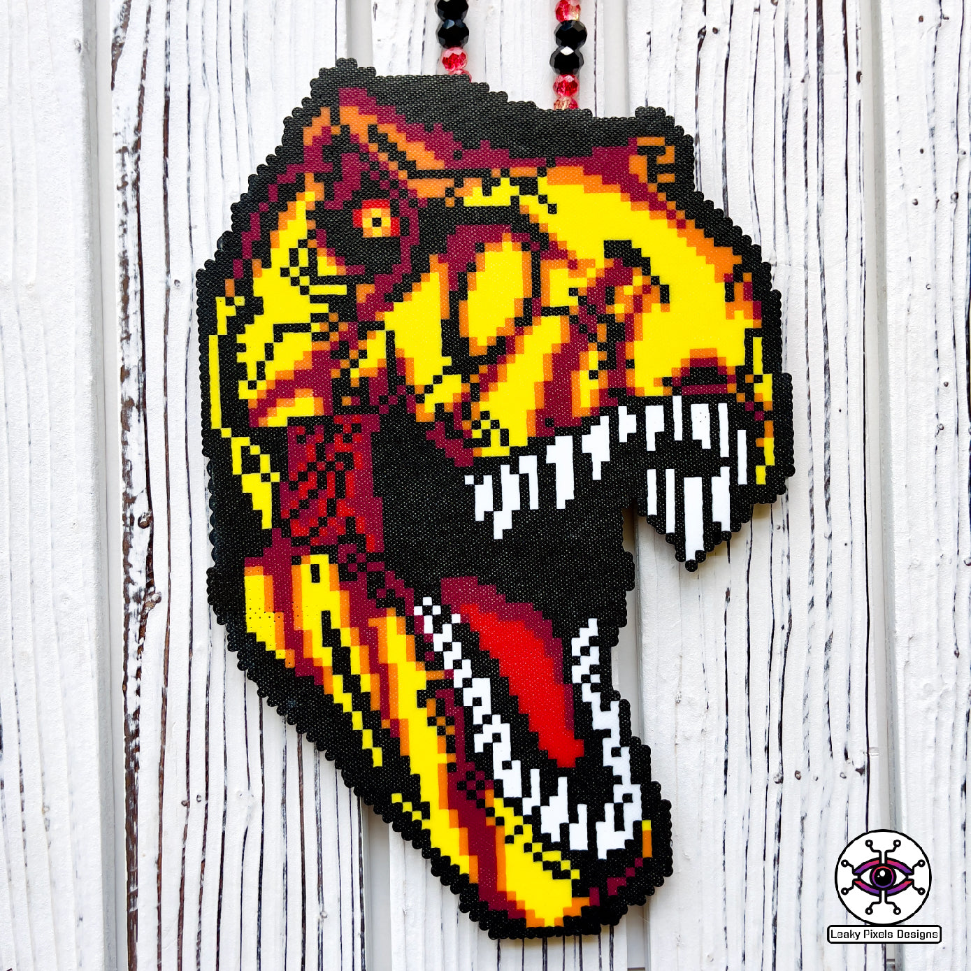 Jurassic Park T-Rex perler necklace made by leaky pixels. T-rex head with mouth open with yellow, red, orange and crannapple perler beads. necklace is made of red glitter and black pony beads.