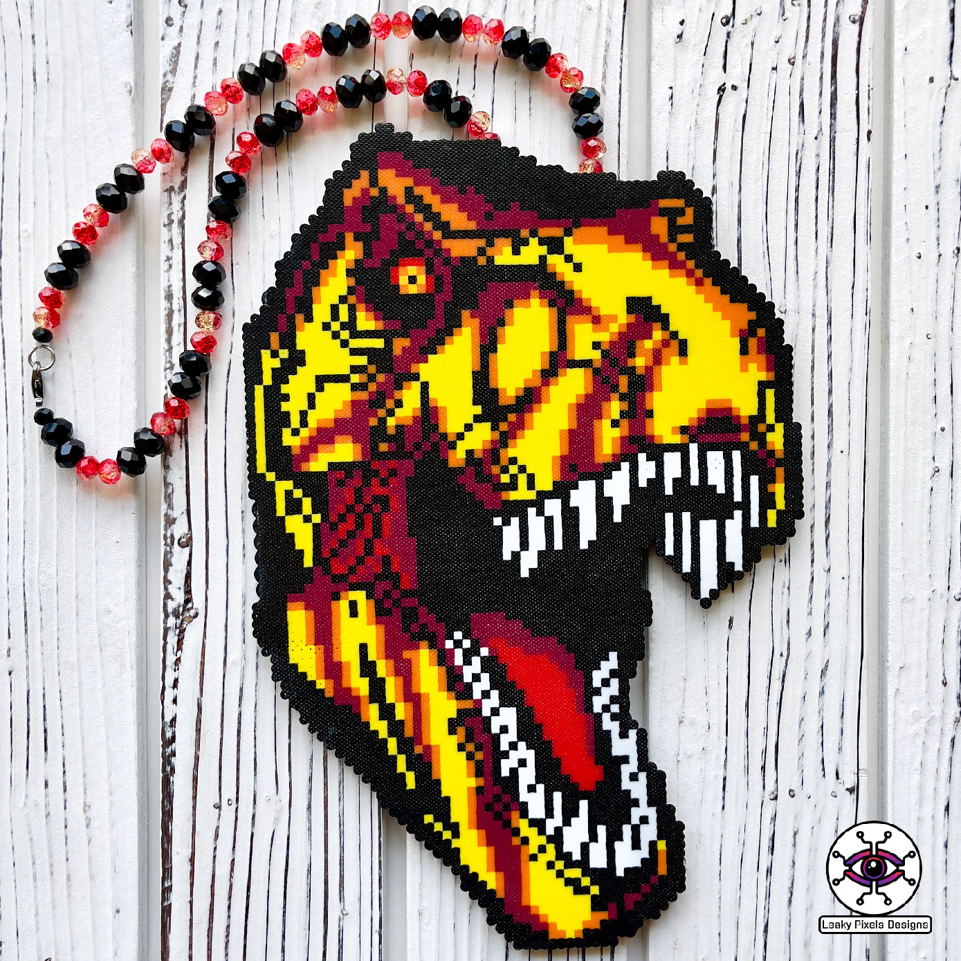 Jurassic Park T-Rex perler necklace made by leaky pixels. T-rex head with mouth open with yellow, red, orange and crannapple perler beads. necklace is made of red glitter and black pony beads.