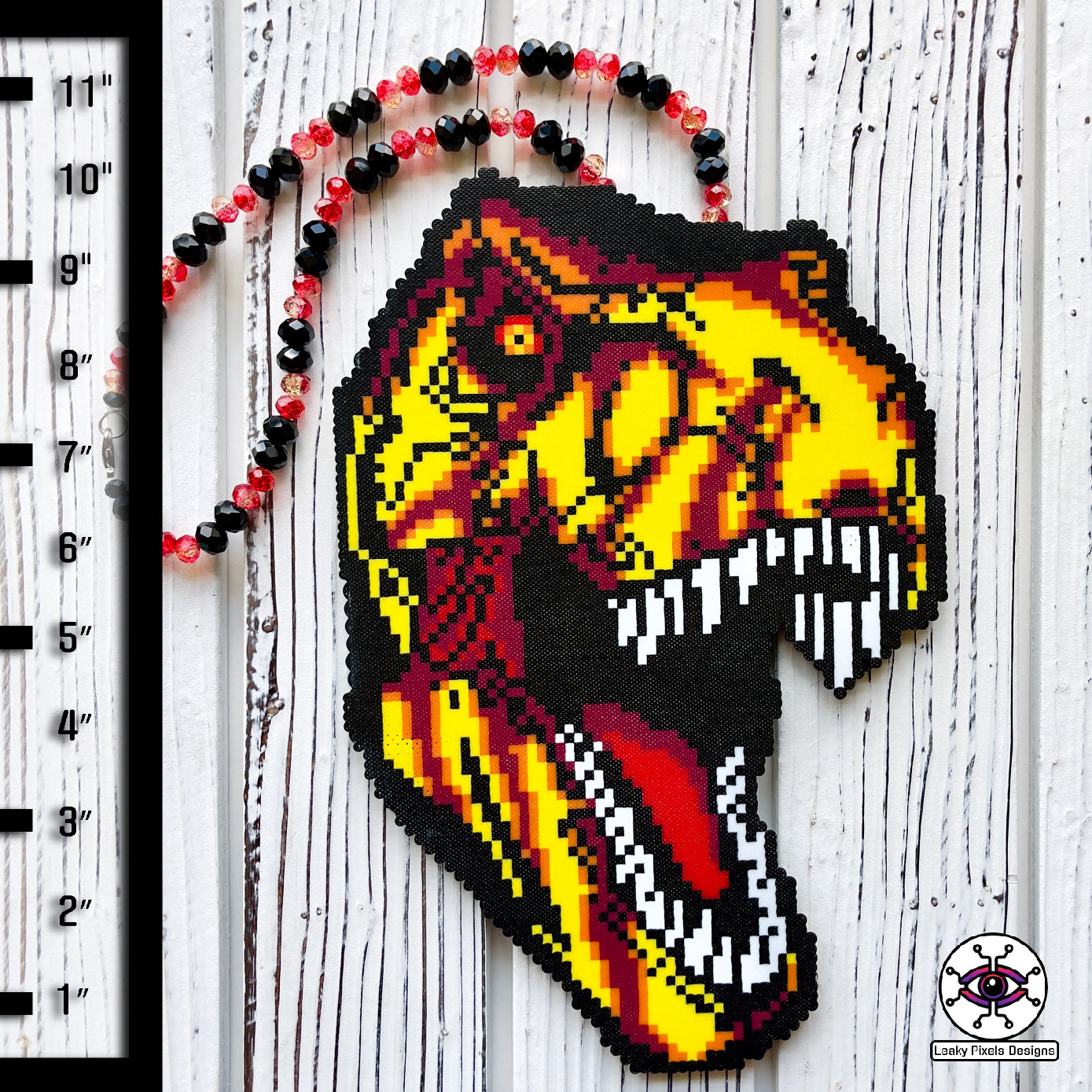 Jurassic Park T-Rex perler necklace made by leaky pixels. T-rex head with mouth open with yellow, red, orange and crannapple perler beads. necklace is made of red glitter and black pony beads.